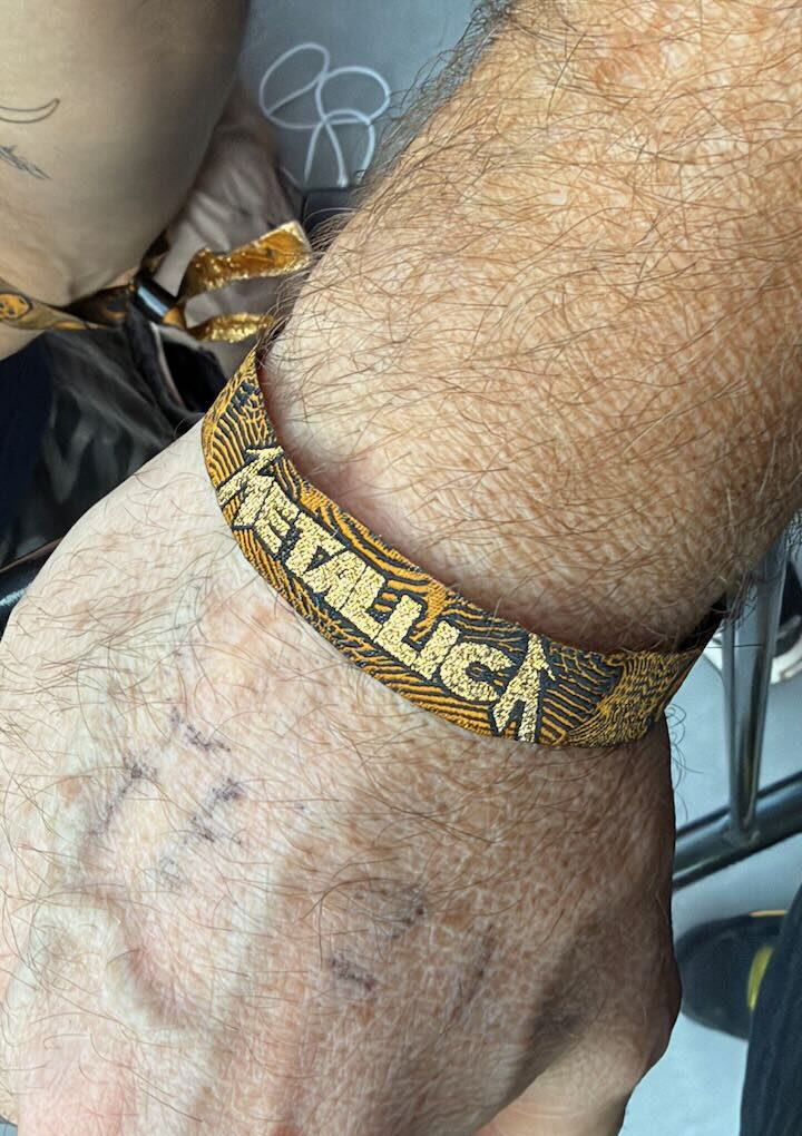 A gold and black Metallica floor wristband on Wesley's wrist, with the faded Sharpie line number still on the back of his hand