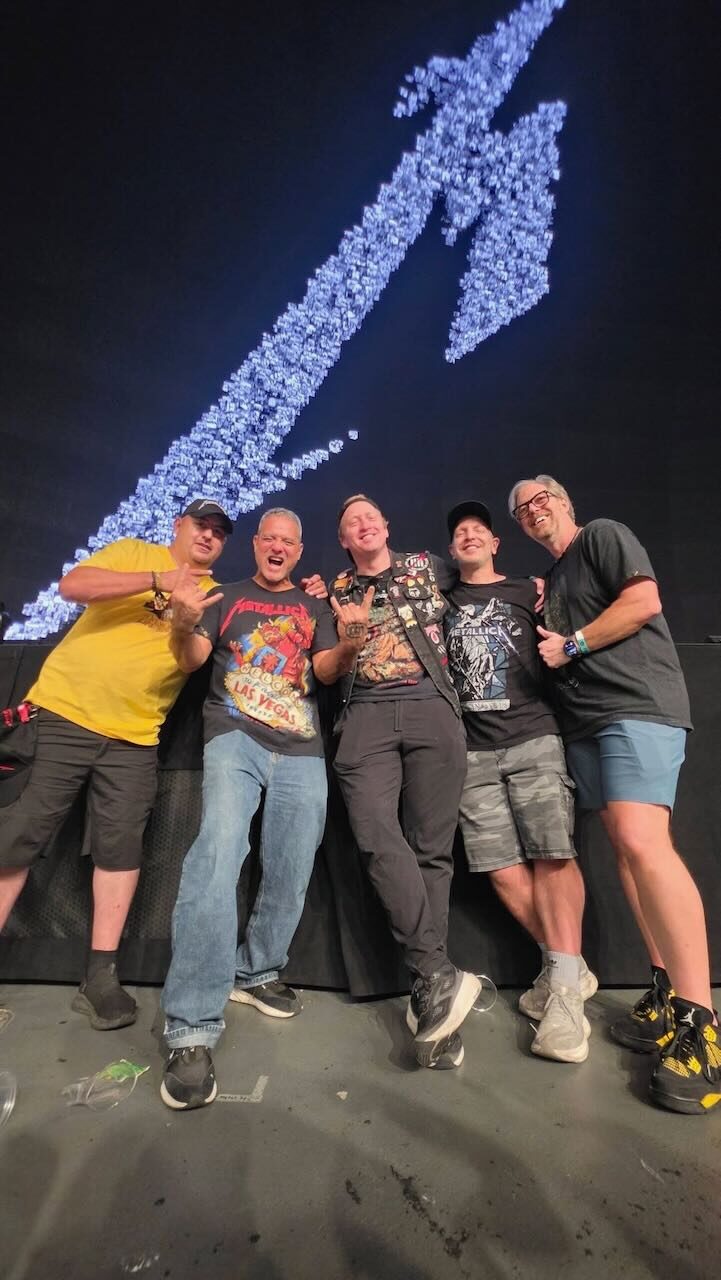 Five people posing under the lit lightning-bolt M Metallica logo after the show, with Wesley on the far right next to Brandon and Brian