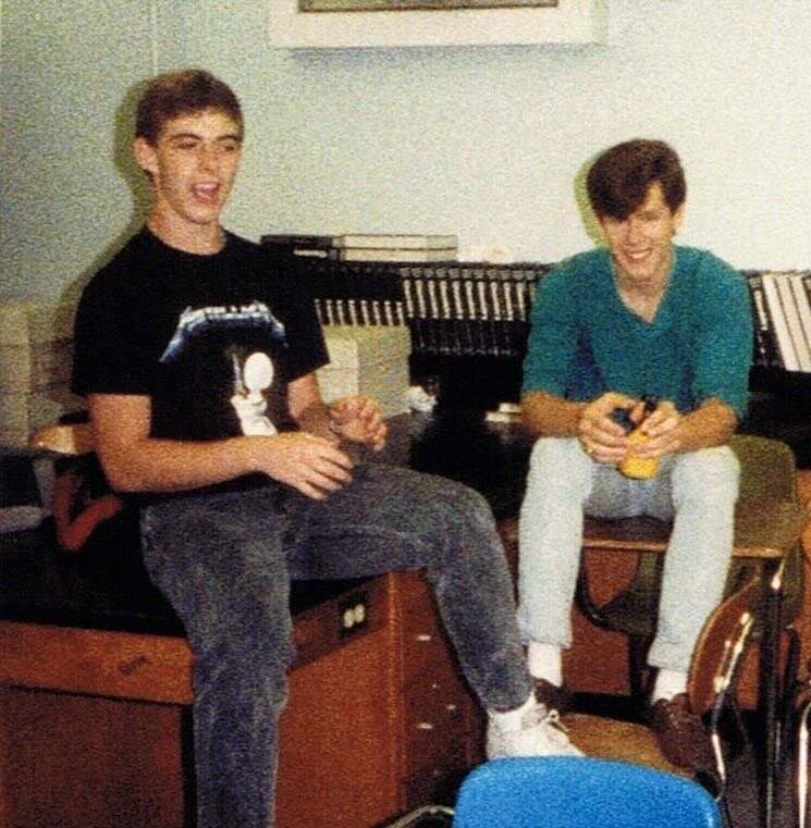 An old high school photo of two teenagers sitting on classroom desks: Wesley on the left, laughing in a black Metallica Metal Up Your Ass shirt, and his friend Marc in a teal shirt