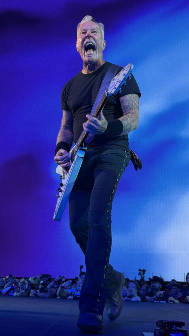 James Hetfield mid-scream at the front of the stage, playing a white Flying V against a blue screen