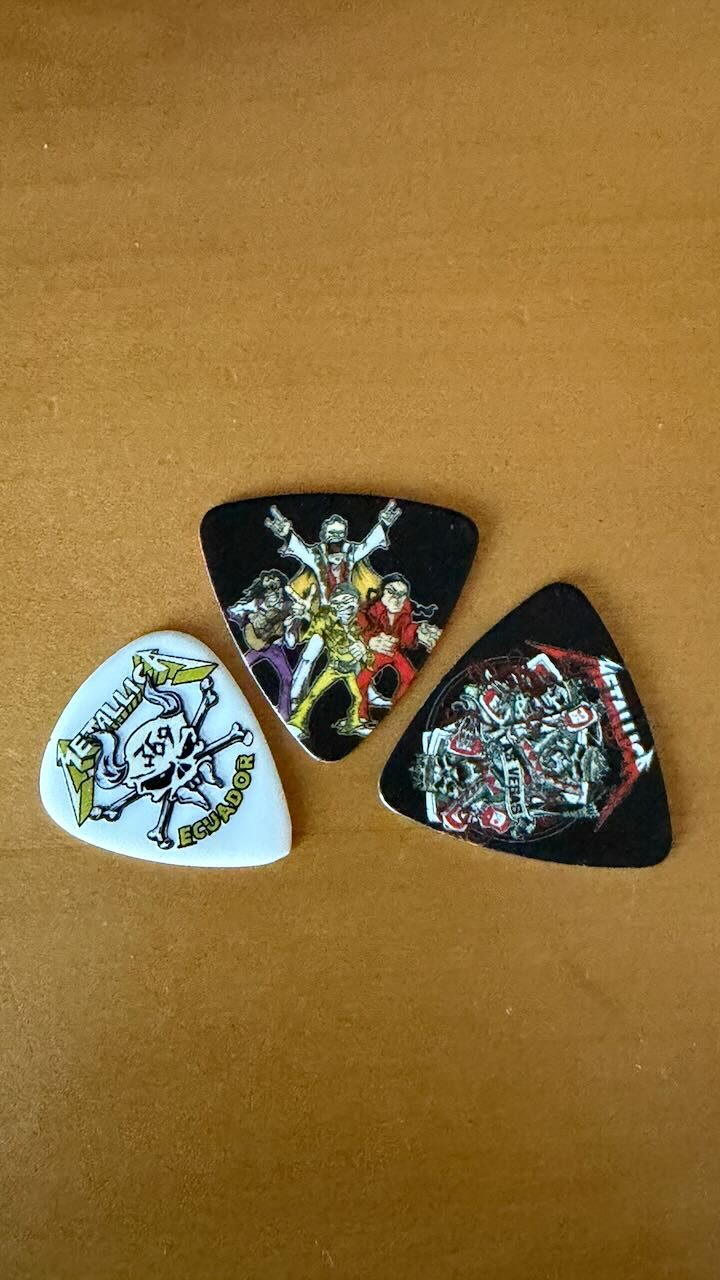 Three Metallica guitar picks on a table: a white Ecuador fan club pick, the black pick Rob Trujillo handed over, and the black blood drive pick